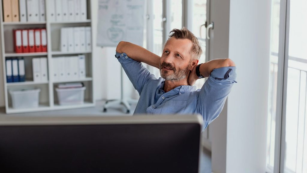 Mann entspannt sitzt am Schreibtisch, Hände hinter dem Kopf, Blick zum Fenster – symbolisiert Pause, Konzentration und Work-Life-Balance