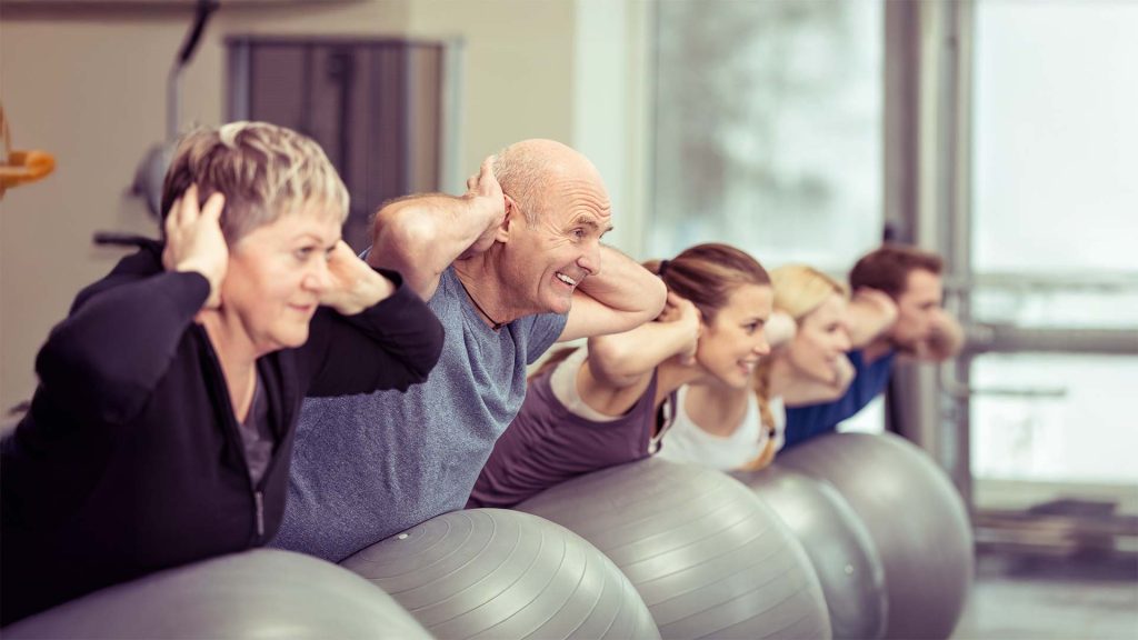 ltere Erwachsene auf großen Gymnastikbällen – Gruppenbild mit mehreren Personen in Fitnessstudio-Umgebung, teilweise mit unscharfen Details rechts im Bild.