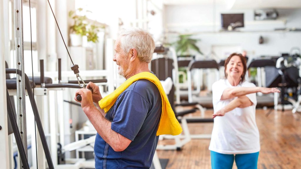 Un homme âgé souriant dans une salle de sport, une serviette jaune autour du cou – arrière-plan flou, appareils de sport et salle d'entraînement