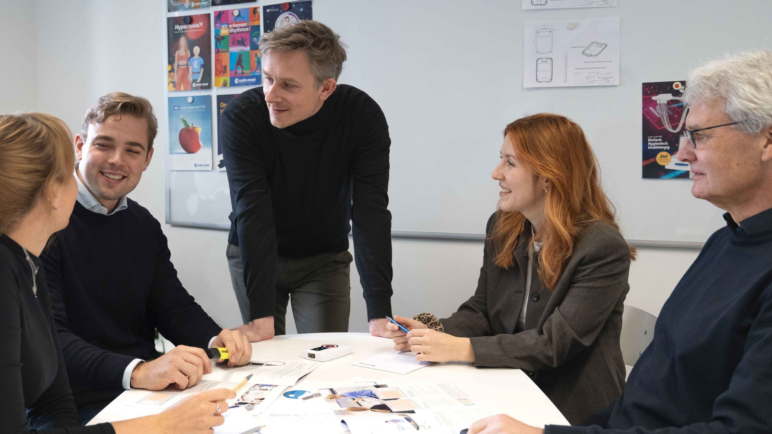 Team-Meeting: Gruppe um Tisch in einem hellen Raum, Poster an der Wand